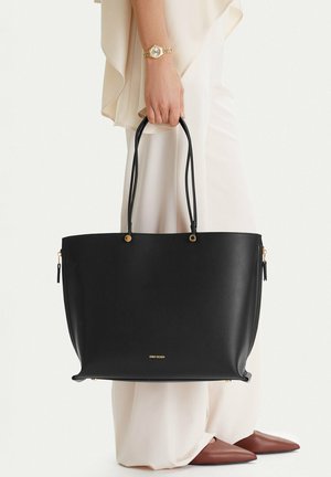 Osoba v bílém oblečení drží velkou černou koženou tašku na dvojitá madla se zlatými kovovými detaily, má na sobě hnědé špičaté boty a zlaté hodinky na zápěstí.