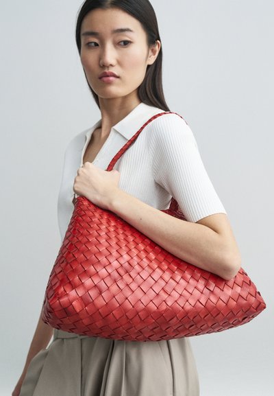 Piros szőtt kézitáska, nagy háromszög alakú, sima bőr textúrával, fonott fogantyúkkal, fehér bordás top és semleges szoknya kíséretében.
