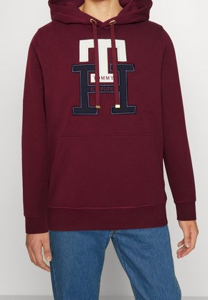 Homme portant un sweat à capuche bordeaux avec un grand patch logo "TH" de Tommy Hilfiger et un jean bleu clair, debout les mains détendues.