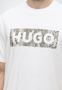 Λευκό T-shirt με υφή και ορθογώνιο μπλοκ με μοτίβο δέρματος φιδιού, που περιέχει τη λέξη "HUGO" με μεγάλα λευκά γράμματα στο στήθος.