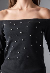 Egy nő fekete, vállvillantós, bordás toppot visel, amelyet az elején és az ujjakon szétszórt apró ezüst szegecsek díszítenek.