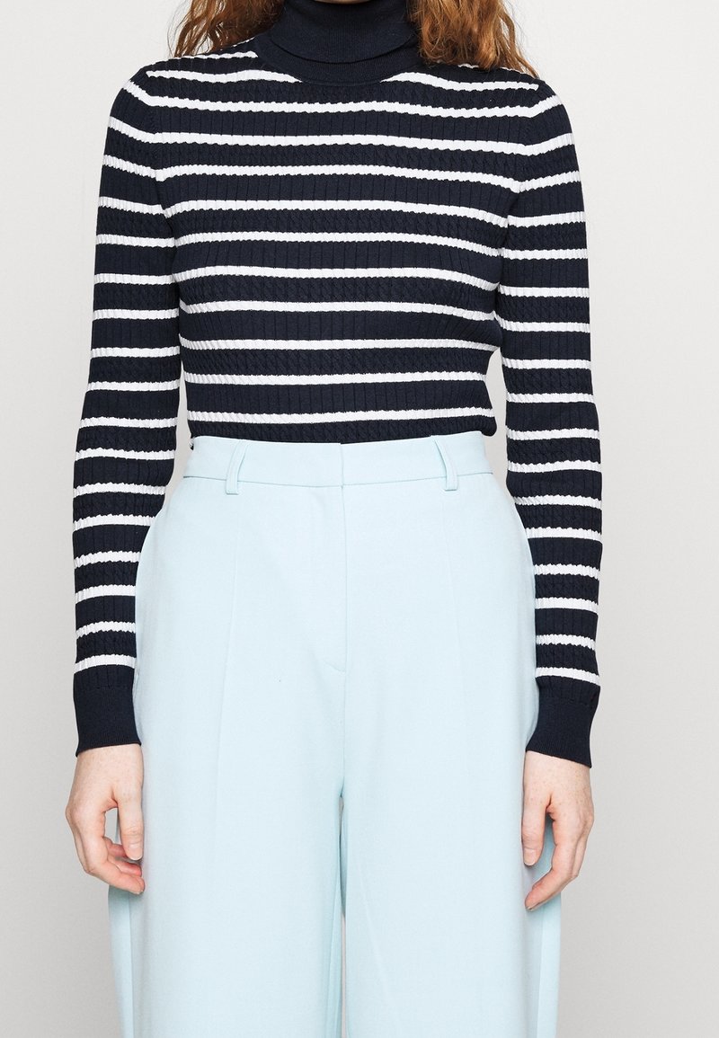 Personne portant un pull à col roulé côtelé rayé noir et blanc et un pantalon taille haute bleu clair sur un fond uni.