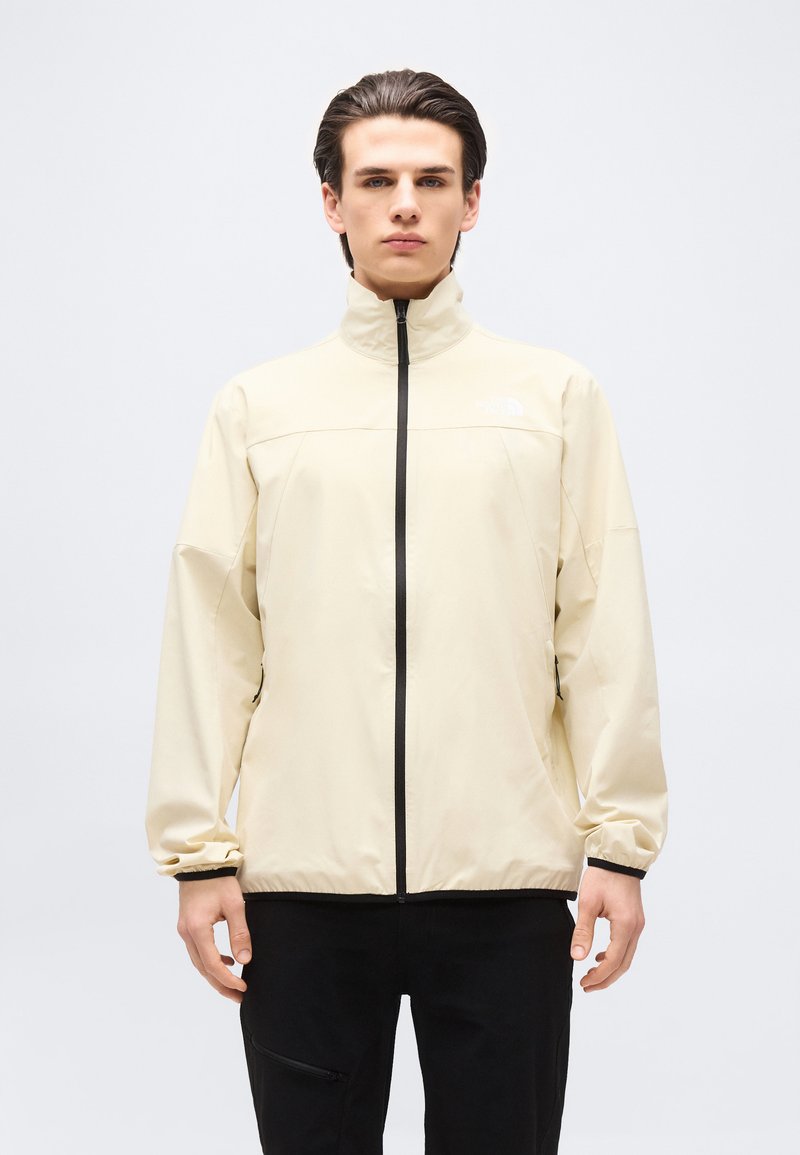 Ung mand iført en beige jakke med lynlås og sorte bukser, stående mod en ensfarvet lys baggrund.