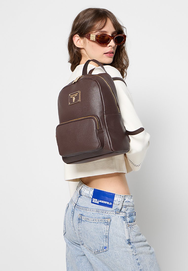 Brauner Leder-Rucksack mit goldfarbener Reißverschluss, Fronttasche und Logo-Plakette. Verfügt über zwei Schultergurte, runde Form und strukturierte Oberfläche.