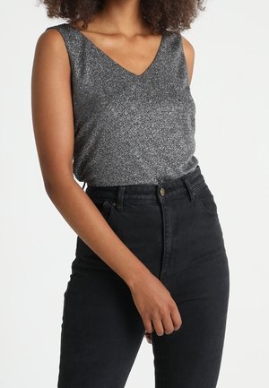 Femme portant un haut gris sans manches et un jean noir taille haute, avec son bras droit détendu et la main près de la taille.