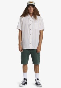 Young man with long curly hair wearing a striped short-sleeve button-up shirt, green shorts, white socks, black high-top sneakers, and a cap.