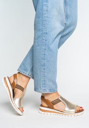 Feet wearing metallic gold and brown slingback sandals with white soles, paired with light blue denim jeans.