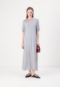 Robe longue gris clair en tissu transparent avec des manches courtes et larges ; associée à des ballerines noires pointues et un sac à main bordeaux.