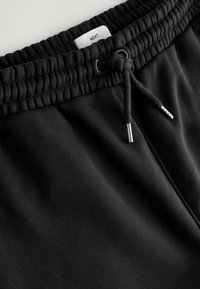 Pantalon de survêtement noir à taille élastique avec cordon de serrage noué, embouts métalliques et étiquette de marque visible indiquant "NEXT" à l'intérieur de la ceinture.