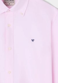 Camisa rosa claro de botones, hecha de una tela suave, que presenta un pequeño logotipo bordado en azul marino en el pecho y un cierre clásico de botones.