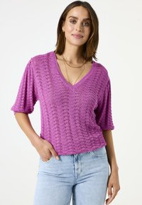 Gehaakte top in felle roze met een V-hals, korte mouwen en een textuurpatroon, gecombineerd met lichtblauwe spijkerbroeken.