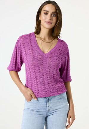 Top en crochet rose vif avec un col en V, des manches courtes et un motif texturé, associé à un jeans en denim bleu clair.