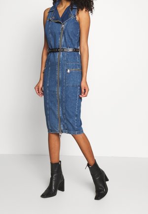 Robe midi sans manches en denim bleu avec fermeture éclair à l'avant, ceinture à la taille, grande poche latérale, portée avec des bottines noires à talons.