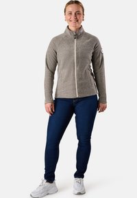 Giacca in pile grigia con zip e colletto alto, caratterizzata da una superficie liscia e chiusura con cerniera, indossata con jeans blu e sneakers bianche.
