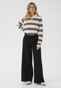Polo rayé en crème et noir ; manches longues, col avec boutons. Pantalons noirs amples et chaussures beiges. Design simple, look décontracté.