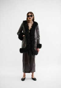 Bruine nepleerjas met zwarte nepbontaccenten, gecombineerd met een bijpassende bruine rok en zwarte flats. Model draagt een donkere zonnebril.
