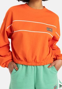 Felpa arancione con collo a girocollo a coste, strisce bianche decorative e orlo elastico aderente. Presenta una grafica colorata sul petto.