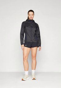 Γυναίκα που στέκεται φορώντας μαύρο φούτερ με φερμουάρ, μαύρες βερμούδες, λευκές κάλτσες και ανοιχτόχρωπα αθλητικά παπούτσια μπροστά σε λευκό φόντο.