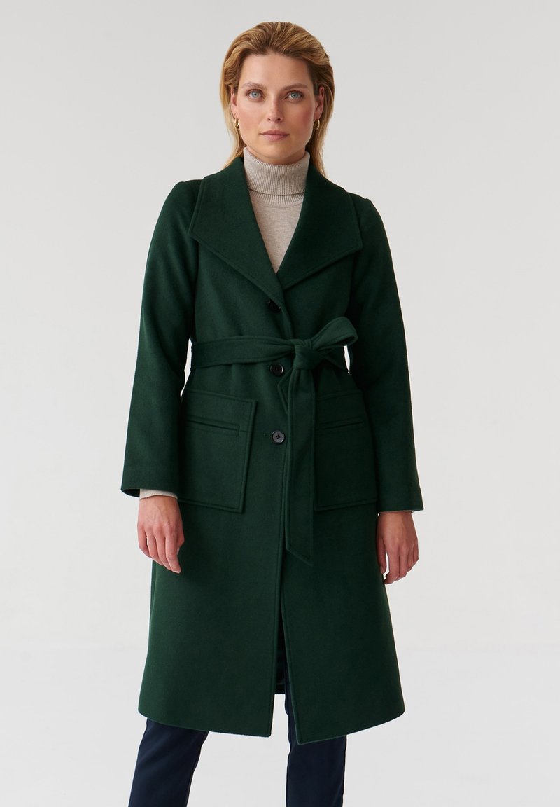 Donna che indossa un lungo cappotto verde scuro con cintura sopra un dolcevita beige e pantaloni blu, in piedi davanti a uno sfondo chiaro e semplice.
