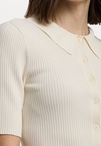 Krémový pletený top s žebrováním, špičatým límcem, krátkými rukávy a předním zapínáním na knoflíky s malými odpovídajícími knoflíky.