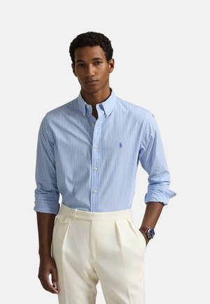 Camisa de botones a rayas de color azul claro y blanco con cuello y mangas largas, que presenta un pequeño logo en el pecho. Combinada con pantalones de color crema.