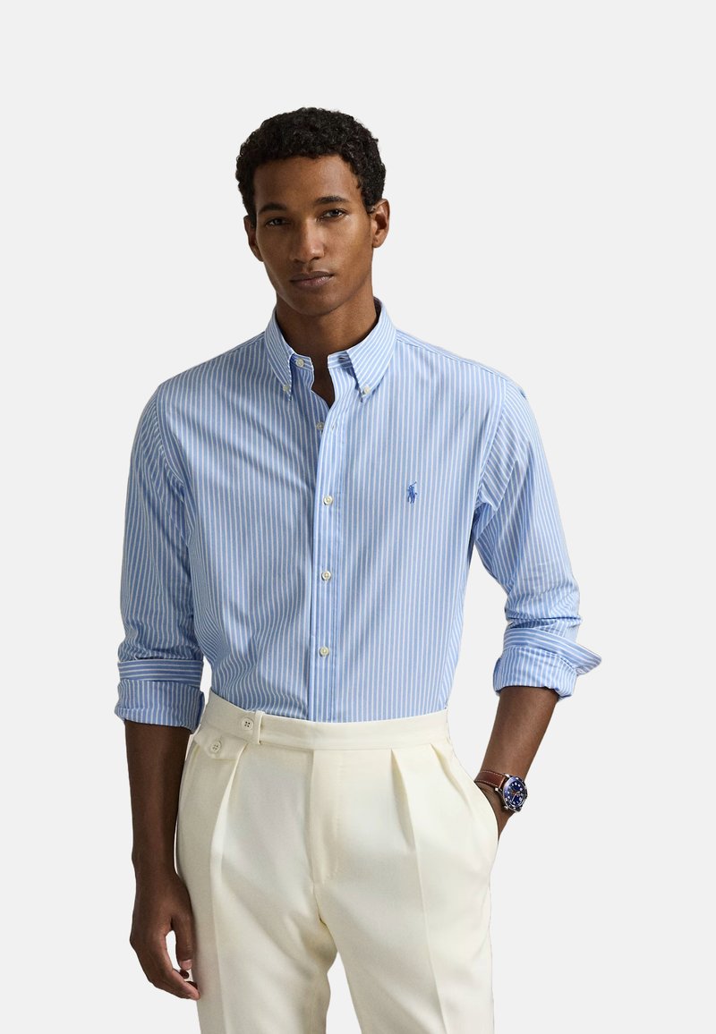 Chemise boutonnée bleu clair et blanche à rayures, avec col et manches longues, ornée d'un petit logo sur la poitrine. Associée à un pantalon crème.