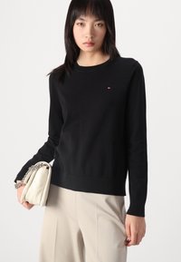Maglione nero a maglia con maniche lunghe e scollo rotondo. Presenta un piccolo logo sul petto. Abbinato a pantaloni beige e una borsa color crema.