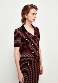 Robe à motif rouge et noir avec un col, des manches courtes, des poches poitrine doubles, des boutons dorés et une silhouette ajustée.