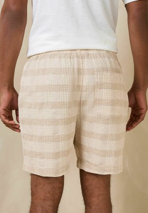 Mann trägt beige-weiße karierte Shorts mit Gesäßtasche, gezeigt von der Taille bis zur Mitte des Oberschenkels, vor neutralem Hintergrund.