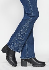 Jambes portant un jean bleu avec une broderie florale sur l'intérieur des mollets et des bottines noires à talons épais sur fond blanc.