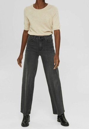 Zwarte jeans met hoge taille en wijde pijpen, voorzien van voorzakken en subtiele stiksels. Gecombineerd met een beige gebreide top met korte mouwen.