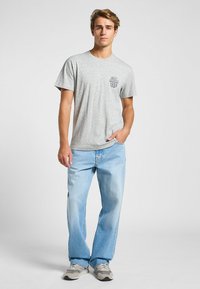 Camiseta de algodón gris de manga corta, con un estampado de logo en el lado izquierdo del pecho; combinada con jeans de mezclilla de pierna recta en azul claro.
