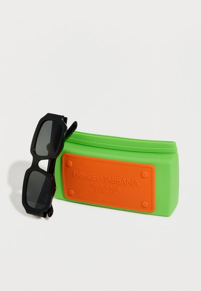 Μαύρα ορθογώνια γυαλιά ηλίου ακουμπισμένα σε μια πράσινη θήκη σιλικόνης Dolce & Gabbana με πορτοκαλί λογότυπο.