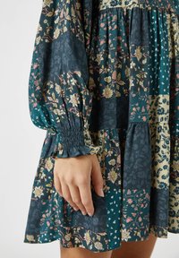 Robe à motif floral avec des manches volantées, un ourlet en plusieurs niveaux et un mélange de couleurs vert foncé, beige et pastel dans diverses textures de textile.