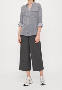 Camicia a righe con bottoni e due tasche sul petto, abbinata a pantaloni a gamba larga grigi e sneakers bianche con dettagli neri. Vestibilità casual.