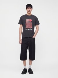 Mannelijk model draagt een zwart T-shirt met een bruine hond die een rode roos vasthoudt en tekst "Who's a good boy?", gecombineerd met zwarte wijde shorts en zwarte schoenen.