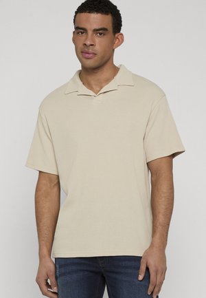 Homme aux cheveux courts et bouclés portant un polo beige à manches courtes et un jean bleu foncé, debout devant un fond clair uni.