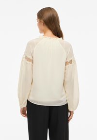 Blusa bege clara com decote com babados, mangas longas transparentes e detalhes em renda nos ombros. Tecido suave, ajuste solto.