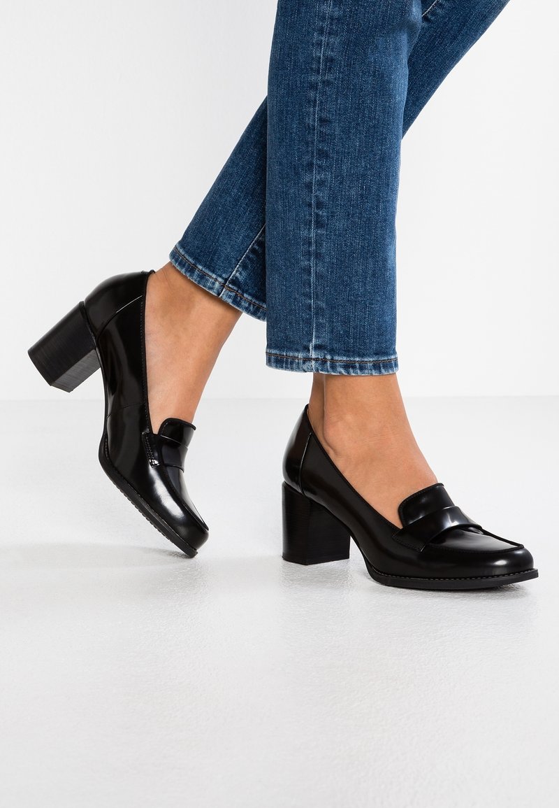 Chaussures à talons en cuir verni noir avec un bout rond et un détail de nœud décoratif. Caractérisées par un talon épais et une semelle texturée.