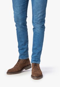 Braune Wildleder-Ankelstiefel mit strukturiertem Finish, runder Spitze und Schnürverschluss, kombiniert mit schmalen blauen Jeans.