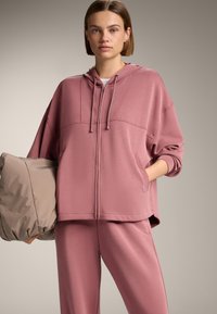 Uvoľnená mikina s kapucňou na zips v tlmenej ružovej farbe s klokanskými vreckami, kombinovaná s ladnými teplákmi, vyrobená z príjemného materiálu a s rebrovanými manžetami.