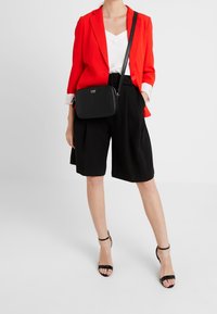 Rote Blazer über einem weißen Oberteil, kombiniert mit schwarzen hochtaillierten Shorts und schwarzen Riemchen-Heels. Eine kleine schwarze Handtasche wird quer über den Körper getragen.