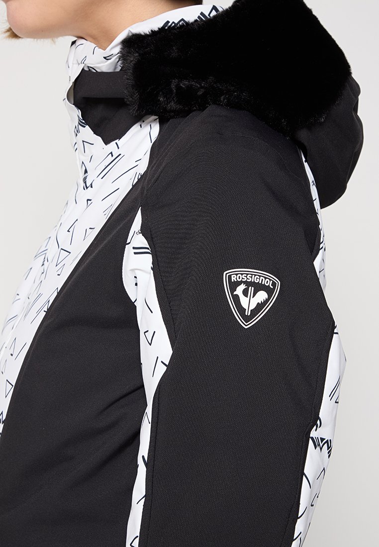 Osoba ubrana w czarną i białą kurtkę Rossignol z kapturem wykończonym sztucznym futrem, pokazująca profil boczny od ramienia do podbródka.