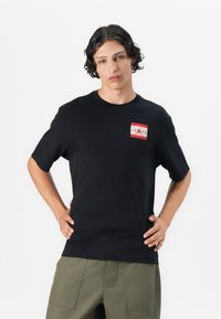 Sort bomulds t-shirt med et rødt og hvidt grafisk logo på venstre bryst. Rund halsudskæring, korte ærmer og almindelig pasform.