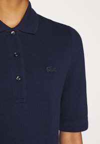 Polo bleu marine avec trois boutons noirs et un logo crocodile brodé ton sur ton sur le côté gauche de la poitrine, porté sur le torse supérieur d'une personne.
