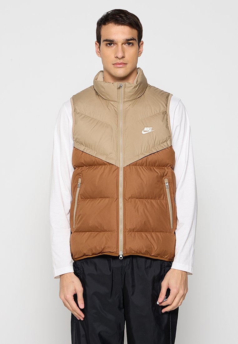 Gilet rembourré beige et marron, avec un col haut, un matelassage horizontal et des poches zippées sur le devant. Logo Nike sur la poitrine.