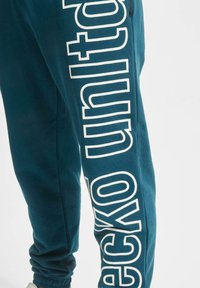 Teal joggebukser laget av mykt stoff har en tilpasset mansjett og stor hvit "ecko united" logo langs venstre ben i fet skrift.