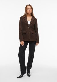 Blazer em camurça castanha com dois botões, lapelas notch e dois bolsos frontais, combinado com calças pretas e saltos pretos pontiagudos.