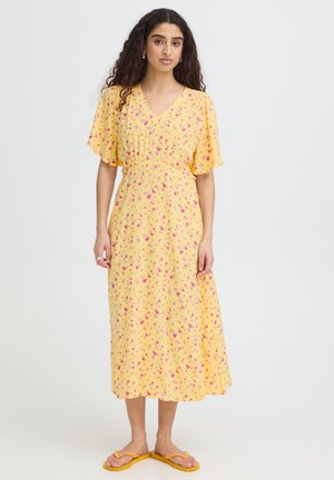 Kobieta z długimi kręconymi włosami ubrana w żółtą sukienkę midi z różowym kwiatowym wzorem i żółte klapki, stojąca na jednolitym tle.