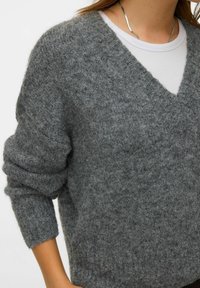 Szary dziergany sweter z dekoltem w serek, długimi rękawami i teksturowaną tkaniną. Nałożony na białą koszulę dla dodatkowego efektu warstwowego.